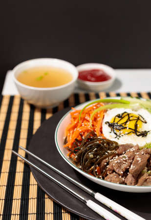 A popular dish of Korean cuisine, Pibimpap. Boiled rice with salads of carrots, kimchi, wood mushrooms and pickled beef. Served with fried egg, broth and sauce.Close-up, on a black background, vertical with copy spaceの写真素材
