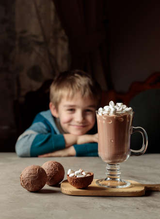 A blond boy with a smile looks at a glass of cocoa or hot chocolate with marshmallows. Chocolate bombs for making cocoa. Selective focusの写真素材