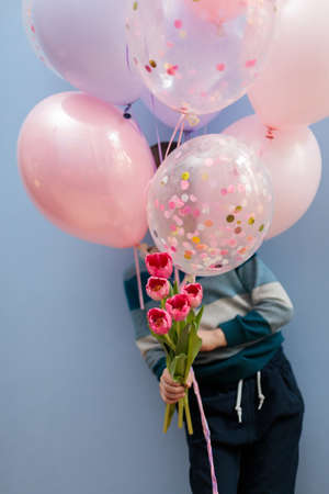 The child holds a bunch of balloons and a bouquet of tulips, the face is hidden behind the balloons. Vertical frame on a blue background.の写真素材