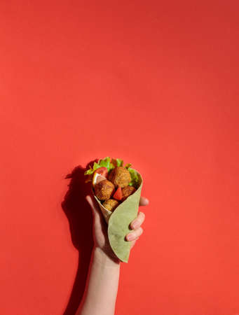 street food. The hand holds a shawarma with falafel, or meatballs. On a red background, hard light. Copy of the spaceの写真素材