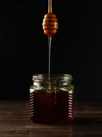 Dark honey, thick syrup or jam flows from a wooden spoon into a jar. Close-up, on a black background, vertical frameの写真素材