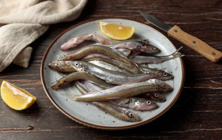 Raw small fish on a plate with a slice of lemon. Close-up on a dark wooden background.の写真素材