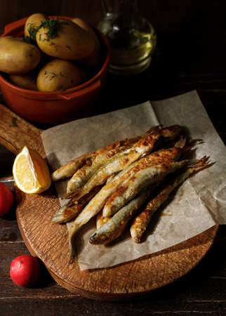 Fried fish on parchment, boiled potatoes and fresh vegetables in the background. Still life, low keyの写真素材
