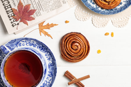 A cup of tea in a vintage cup, cinnamon and raisin buns and a newspaper on a light wooden...
