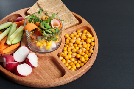 Wooden tray with dividers, with pieces of fresh vegetables, chickpeas and grain loaves. On a black background. Horizontal photo with space for text.の写真素材
