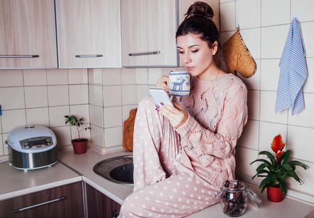 Young woman in pajamas having breakfast in the kitchen while using her phoneの写真素材