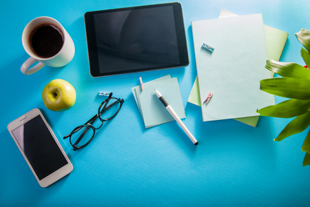Workspace of a creative office employee. Working with a cup of coffee, using a phone and tablet on blue backgroundの写真素材