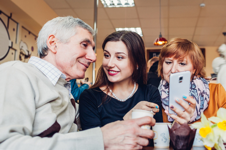 Happy family spending time together. Senior family couple with adult daughter using a smartphone in the cafe drinking coffee.の写真素材