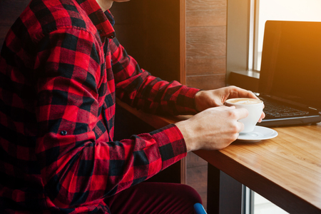 Young man freelancer enjoys coffee in cafe using laptop. Self-employed worker drinking coffee and waiting for a client by the window.の写真素材
