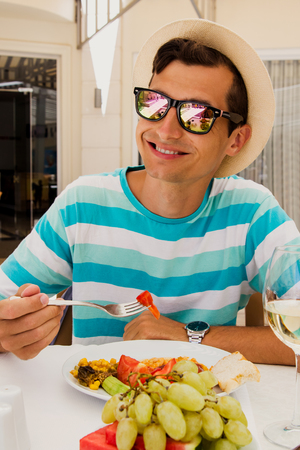 Happy young man eating food in hotel restaurant. All inclusive concept. Summer vacation. Having lunch with glass of wine and fruits.の写真素材