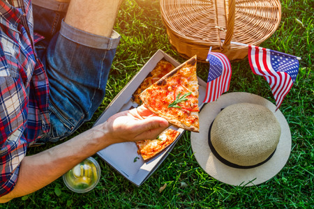 Young man celebrating Independence Day of America in the park. Man and woman having pizza with lemonade. Picknick basket with USA flag.の写真素材