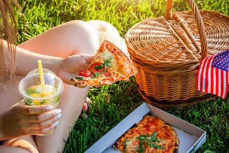Young woman celebrating Independence Day of America in the park. Man and woman having pizza with lemonade. Picknick basket with USA flag.の写真素材