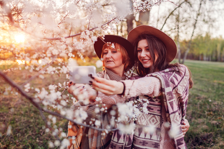 Happy middle-aged mother and her daughter hugging and taking selfie in blooming spring garden at sunset. Mother's day concept. Family valuesの写真素材