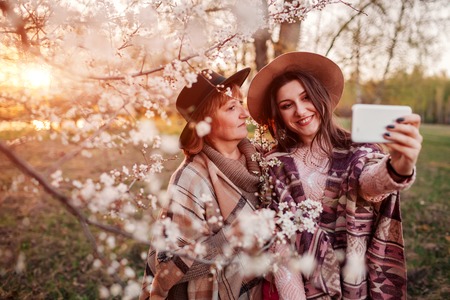 Happy middle-aged mother and her daughter hugging and taking selfie in blooming spring garden at sunset. Mother's day concept. Family valuesの写真素材