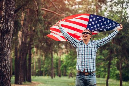 Happy man running with USA flag in park at sunset. Celebrating Independence Day of America. July 4th. Man having funの写真素材