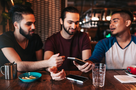 Group of mixed race young men using phone and talking in lounge bar. Middle-eastern friends having fun in cafe drinking coffeeの写真素材