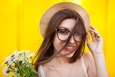 Happy hipster girl wearing glasses and hat with flowers against yellow background. Summer outfit. CLose up of smiling womanの写真素材