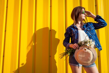 Portrait of young hipster girl wearing glasses and hat with flowers against yellow background. Summer outfit. Fashion. Space.の写真素材