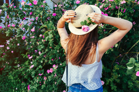 Young hipster girl wearing hat walking by blooming roses. Woman enjoys flowers in park. Summer outfit. Fashionの写真素材