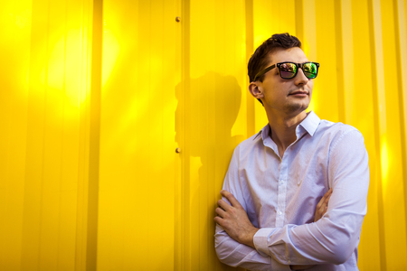 Close up of young stylish man in white shirt and sunglasses standing against yellow wall outdoors. Summer fashionable outfit.の写真素材