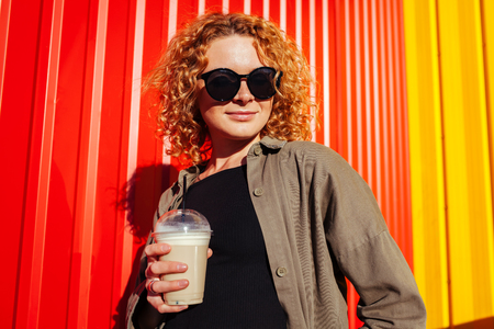 Pretty young woman with curly red hair holding coffee against red wall. Stylish summer girl in sunglasses smiling and relaxing outdoorsの写真素材