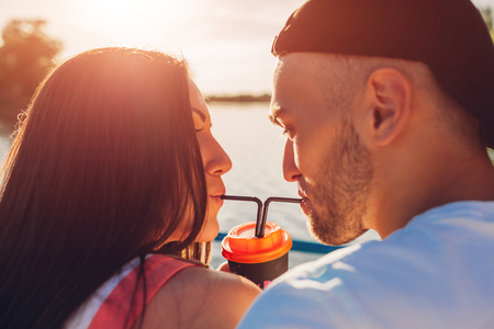 Happy couple in love drinking coffee together with a straw by the river at sunset. Romantic date of young studentsの写真素材