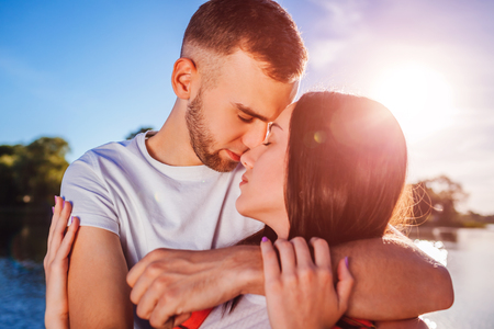 Happy couple in love hugging and kissing by the river at sunset. Romantic date of young peopleの写真素材