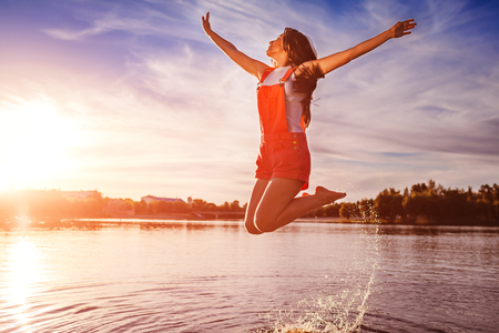 Happy and free young woman jumping and raising arms on river bank. Freedom. Active lifestyle. Summer holidaysの写真素材