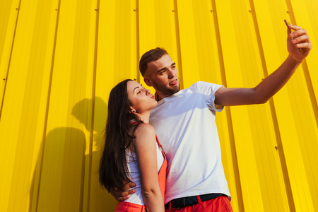 Happy couple in love hugging and taking selfie against yellow wall. Young hipsters having funの写真素材