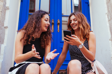 Two best female friends chatting while using smartphone in city. Happy girls talking and laughing sitting by cafe windowの写真素材
