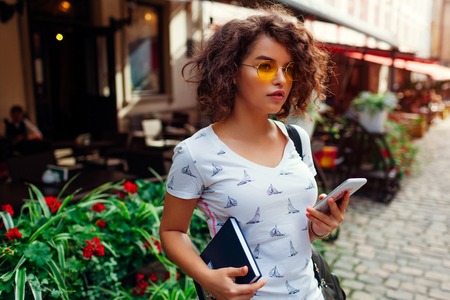Beautiful college girl using smart phone and holding book outdoors after classes. Stylish studentの写真素材