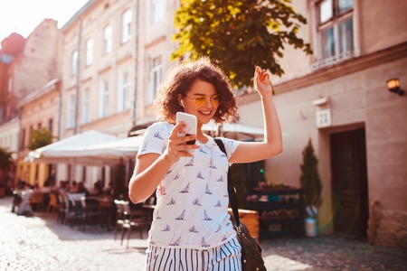 Beautiful young girl using smart phone and listening to the music walking on street. Happy woman dancing and having funの写真素材