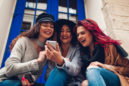 Outdoor shot of three young women looking at smartphone and laughing by cafe. Girls talking and having fun on city streetの写真素材