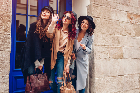 Outdoor shot of three stylish young women looking into the distance on city street. Friends laughing and having fun. Girls waving at somebodyの写真素材