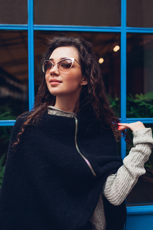 Stylish young woman posing against blue window outdoors. Fashionable outfit. Beautiful model with curly hair smilingの写真素材