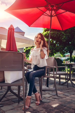 Beautiful stylish young woman with shopping bags chilling in outdoor cafe on city street and talking on phone. Happy girl drinking coffee in summer cafeの写真素材