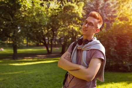 Handsome college guy chilling in spring campus park. Happy man student wears earphones and glasses and looks at cameraの写真素材