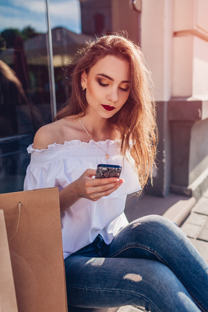 Beautiful stylish young woman with shopping bags chilling on city street and using smartphone. Happy sitting by showcase outdoorsの写真素材