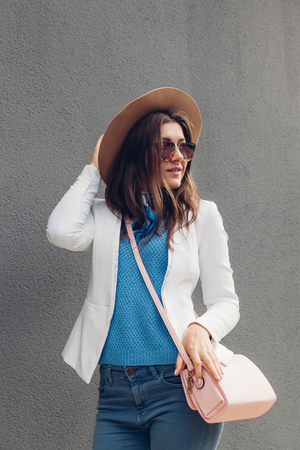 Fashion portrait of pretty young woman wearing jacket, hat, sunglasses and holding bag over grey background. Summer outfitの写真素材