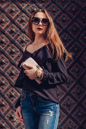 Outdoor portrait of a beautiful stylish woman in sunglasses on city street. Fashion model wearing summer clothing and accessories. Lady holding a purseの写真素材