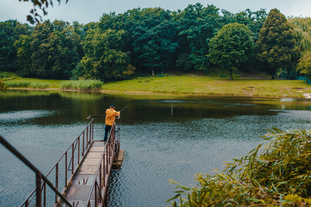 Fisherman in yellow jacket standing on pier of the lake and fishing on rainy dayの写真素材
