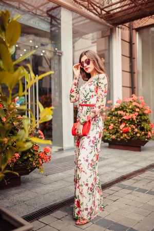Young woman wearing classic dress with accessories outdoors. Beautiful lady poses by modern building in city. Fashionの写真素材