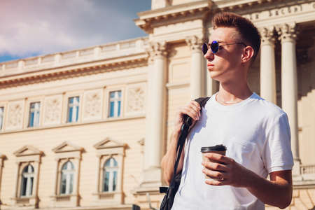 Handsome college man drinking coffee against the university. Guy student with backpack relaxing after classesの写真素材