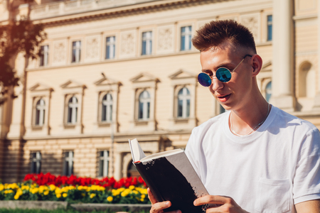 Handsome college man reading a book sitting by university. Happy guy student with backpack learning outdoors. Educationの写真素材