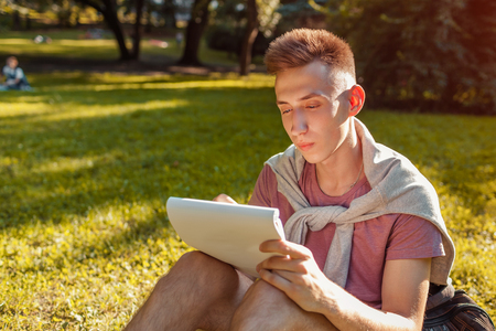 Handsome college man writing notes in copybook in campus park. Happy guy student learning outdoors sitting on grassの写真素材