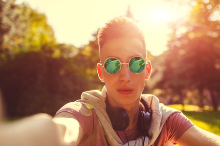 Handsome college man chilling in spring campus park. Happy guy student in sunglasses takes selfieの写真素材