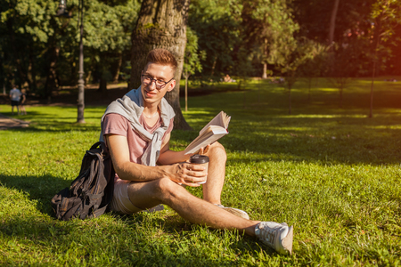 Handsome college man reading a book and drinking coffee in campus park. Happy guy student learning sitting on grass.の写真素材