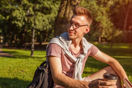 Handsome college man reading a book and drinking coffee in campus park. Happy guy student learning sitting on grass.の写真素材