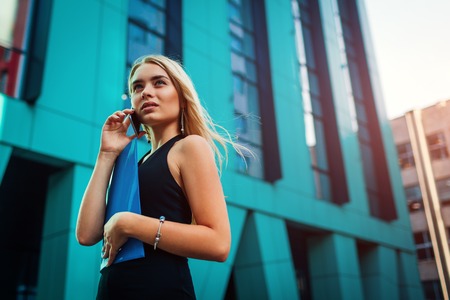 Young blonde businesswoman calling on smartphone and holding folder by business center in city. Communiationの写真素材