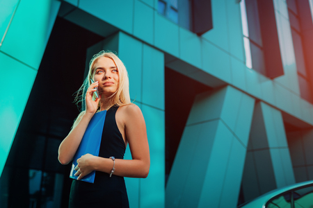 Young blonde businesswoman calling on smartphone and holding folder by business center in city. Communiationの写真素材